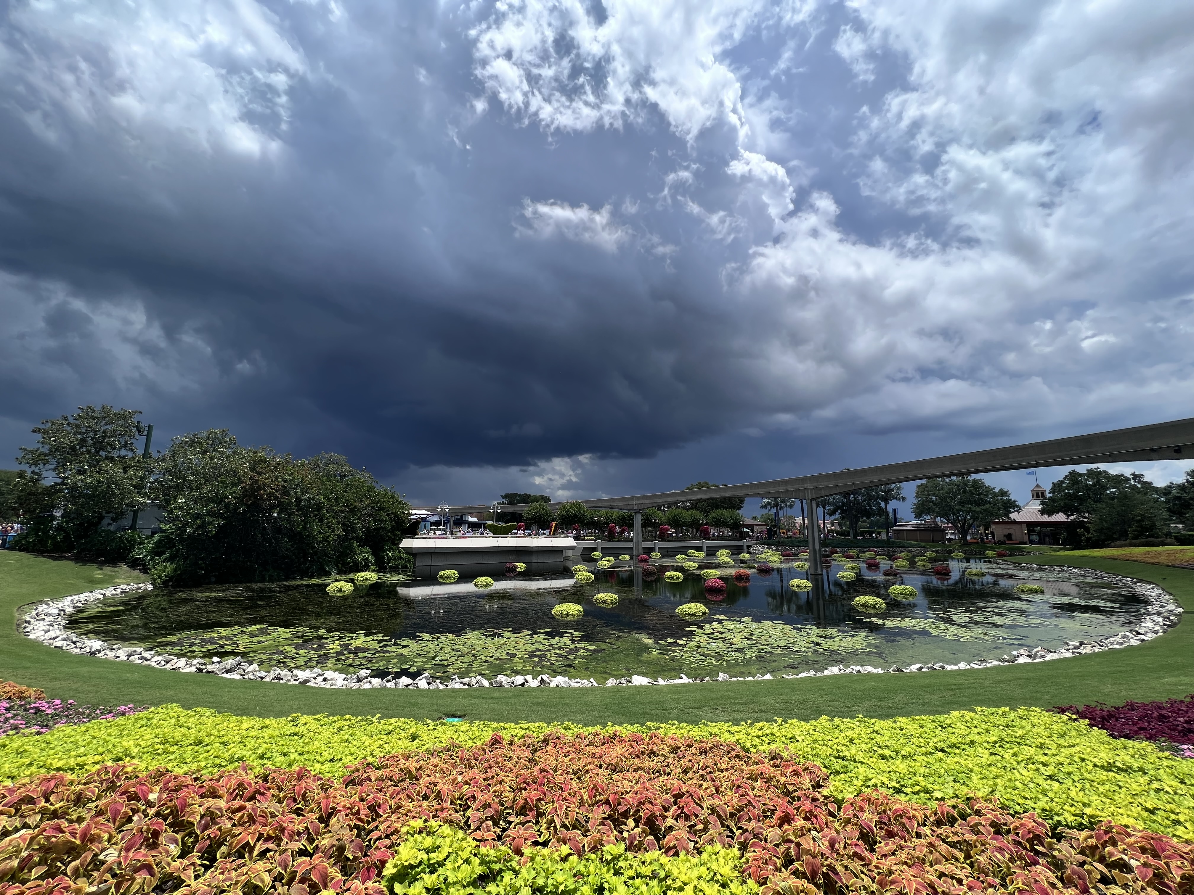 Dramatic storm clouds building over EPCOT's monorail and Flower and Garden Festival pond, with colorful plantings in the foreground.