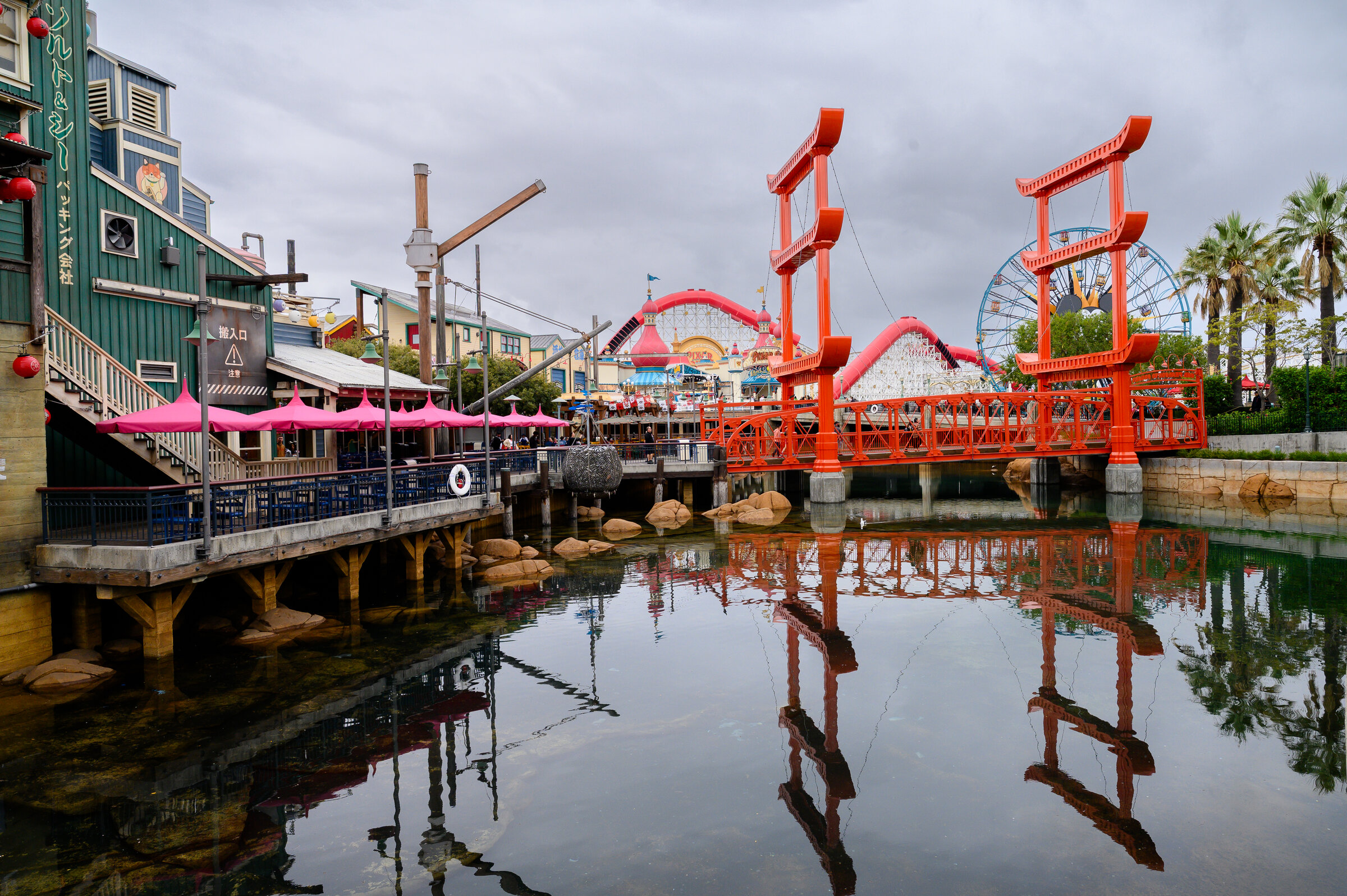 Pixar Pier at Disney California Adventure: the bright red entry gates over Paradise Bay with Incredicoaster's track loops and the Pixar Pal-A-Round Ferris wheel reflected in the water under cloudy skies