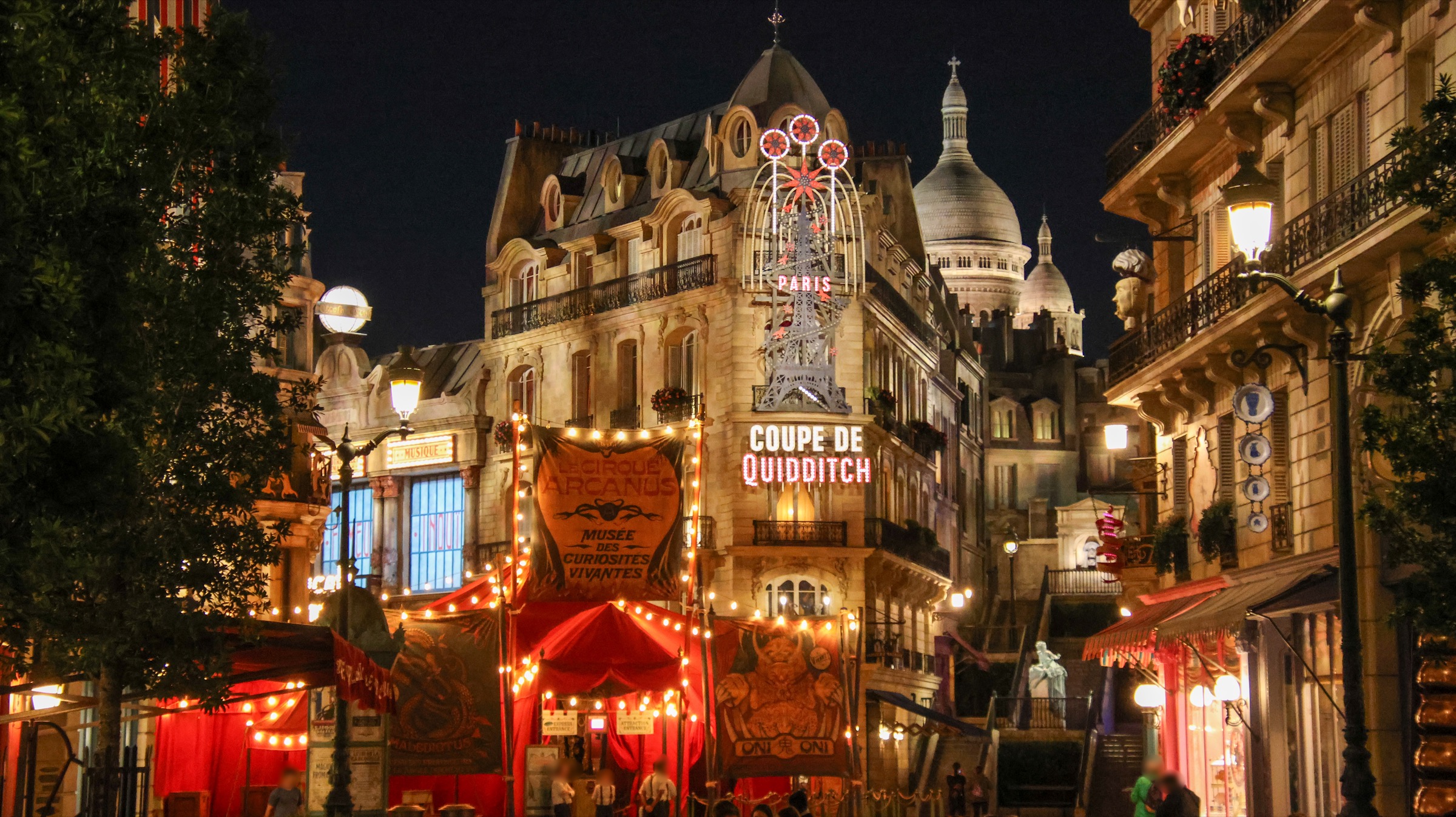 The Wizarding World of Harry Potter &ndash; Ministry of Magic at night: Haussmann-style Parisian buildings glow with warm light, string lights arc over cobblestone streets, and the turrets of a replica Sacr&eacute;-C&oelig;ur rise against the dark sky